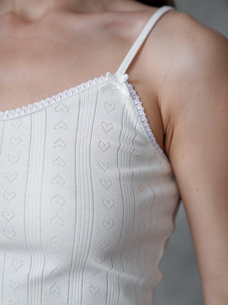 Puede incluir: Camisola blanca con recortes en forma de coraz&oacute;n y tirantes finos. La parte superior presenta una textura acanalada y un delicado ribete de encaje a lo largo del escote y los tirantes. Un peque&ntilde;o lazo adorna la correa. La prenda es de color claro y neutro.