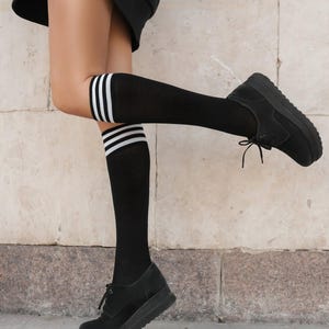 Könnte beinhalten: Schwarze kniehohe Socken mit drei weißen Streifen am oberen Rand, kombiniert mit schwarzen Plateauschuhen. Die Schuhe haben Schnürsenkel und eine Wildleder-ähnliche Textur. Das Outfit befindet sich vor einer neutralen, strukturierten Wand.