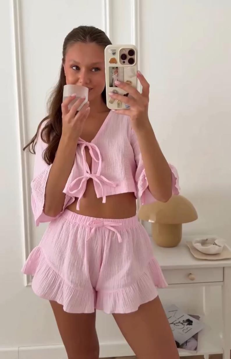 May include: A light pink two-piece set consisting of a cropped top with three-quarter length sleeves, a V-neckline with two front tie closures, and matching high-waisted shorts featuring a ruffled hemline and a drawstring waist with a tie detail.
