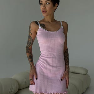 Puede incluir: Un vestido lencero rosa claro con ribete de encaje blanco y tirantes finos. El vestido tiene una textura acanalada con un sutil patr&oacute;n de diamantes y un dobladillo festoneado. El vestido es de largo mini.