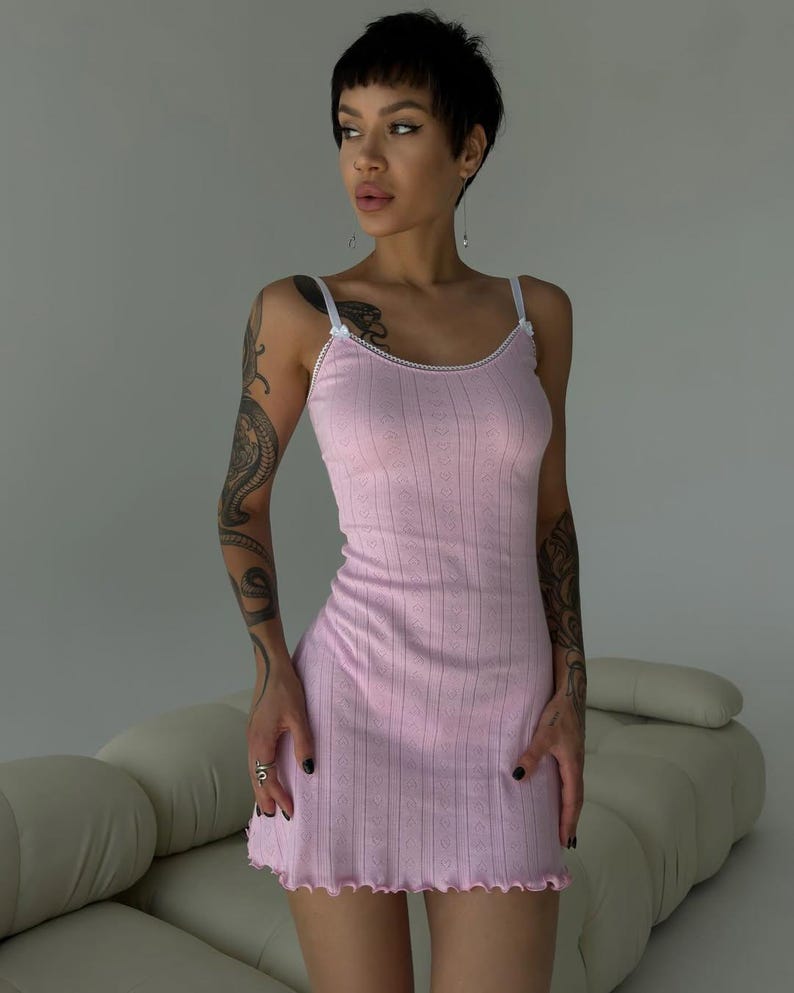 Puede incluir: Un vestido lencero rosa claro con ribete de encaje blanco y tirantes finos. El vestido tiene una textura acanalada con un sutil patr&oacute;n de diamantes y un dobladillo festoneado. El vestido es de largo mini.