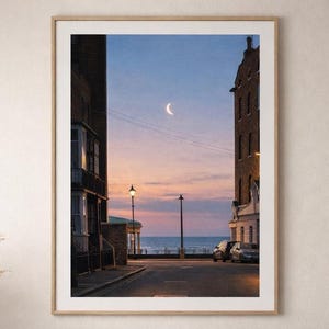 Puede incluir: Impresión enmarcada de una escena de calle costera al anochecer. La imagen presenta una luna creciente en un cielo azul crepuscular, con edificios que bordean la calle y el océano visible en la distancia. La impresión está en un marco de madera.