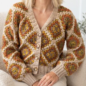 May include: A long-sleeve cardigan constructed from crocheted granny squares in shades of orange, yellow, olive green, and brown. The garment features a V-neckline, a front button closure with four marbled buttons, and ribbed knit cuffs and hem.