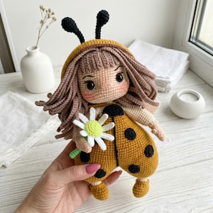 May include: A handmade crochet doll in a ladybug costume. The doll has a yellow body with black spots, brown hair, and a daisy. The doll's face is detailed with stitched features and rosy cheeks. The doll is holding a white and green flower.