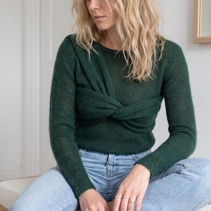 May include: A long-sleeved, dark forest green sweater featuring a round neckline and a prominent twisted knot detail across the chest. The garment is crafted from a fine, slightly sheer knit fabric and is paired with light blue denim jeans.