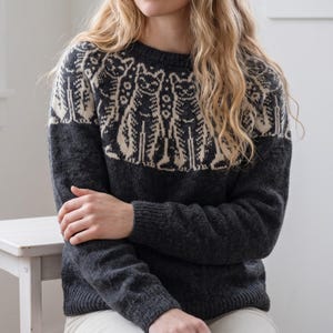 May include: A dark charcoal gray long-sleeve sweater featuring a cream-colored circular yoke pattern. The design displays a repeating motif of stylized cats arranged around the neckline, with ribbed cuffs, a ribbed hem, and a crew neckline.