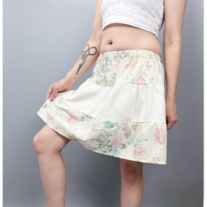 May include: A tiered skirt with a floral pattern and elasticated waistband. The skirt features a cream fabric with a floral print of pink roses and green leaves. The middle tier is a solid cream colour.