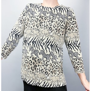 May include: A long-sleeved top with a distinctive animal print. The design incorporates zebra stripes, leopard spots, and stylized animal figures in beige, black, and brown. The top has a round neckline and is made of a soft material.