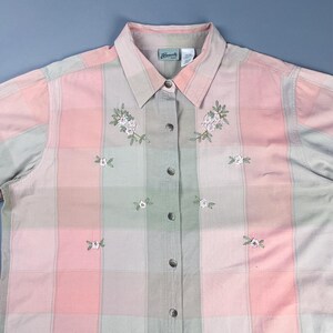 May include: A pink and green plaid button-down shirt with embroidered flowers. The shirt has a collar and long sleeves.