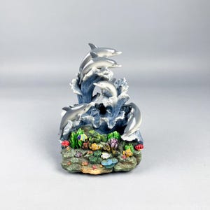 May include: A decorative figurine featuring four silver dolphins leaping from blue waves. The base depicts a colorful coral reef with various fish and aquatic plants. A detailed ocean-themed sculpture.