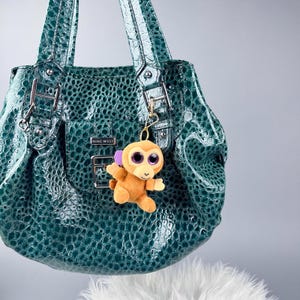 May include: A teal green Nine West handbag with a textured, crocodile-like pattern. A small, plush monkey keychain with large purple eyes and a smiling face hangs from the bag's handle. The bag has silver hardware.