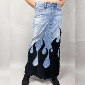 May include: A long, light blue denim skirt featuring a black flame design at the hem. The flames are crafted from a dark, velvety fabric. The skirt has a classic five-pocket design and is a maxi style.