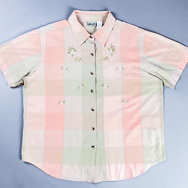 May include: A pink and green plaid button-down shirt with short sleeves and embroidered floral details. The shirt has a collared neckline and a button closure. The brand name "Cherokee" is visible on the label.