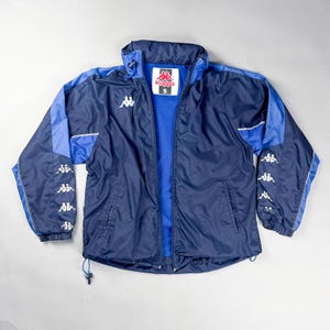 May include: A navy blue and light blue Kappa jacket. The jacket features a full zip, a high collar, and white Kappa logos on the sleeves and chest. The jacket has a size "S" tag.
