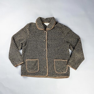 May include: A vintage button-down jacket in a black and tan herringbone pattern. The jacket features a rounded collar, two front pockets, and tan trim. The jacket is made of a textured fabric.