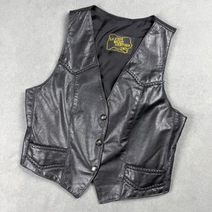 May include: A black leather waistcoat featuring a V-neckline, three front buttons, and braided leather trim detailing on the chest and pockets. The interior is lined with black fabric and includes a label reading LEATHER NIKO LEATHER INT.
