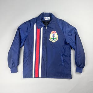 May include: A navy blue jacket with a zip, white and red vertical stripes, and a chest patch with text and a graphic. The jacket has a collared neckline and long sleeves. The jacket is a classic style.