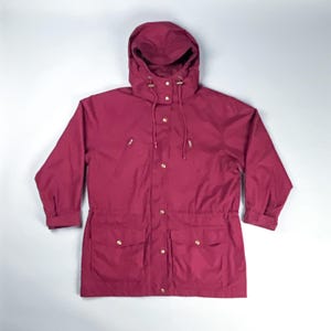 May include: A burgundy hooded jacket with a drawstring hood and gold-coloured buttons and zips. The jacket has two front pockets and a drawstring waist. The sleeves are cuffed at the wrists.