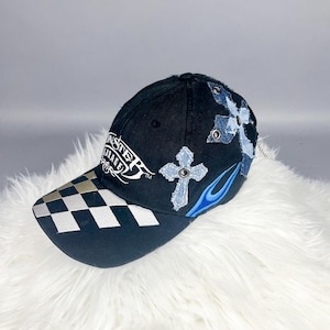 May include: Black baseball cap with a checkered brim and embroidered details. Features a cross design and the text "Monster Garage". Accented with blue flames.