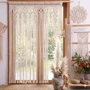 Peut inclure: Rideau en macramé crème avec des motifs géométriques complexes et de longues franges nouées. Le rideau est suspendu devant une fenêtre, avec une tenture murale en macramé assortie et d'autres éléments décoratifs.