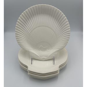 May include: A stack of five off-white ceramic plates shaped like large scallop shells. Each plate features a textured surface with radiating ridges that mimic the natural pattern of a seashell, with a smooth, curved edge and a defined hinge area.
