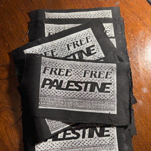 May include: A stack of black fabric patches with the words "FREE PALESTINE" printed in white. The patches have a rectangular shape with a decorative border design. The text is centered on the patch.