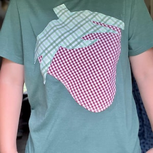May include: A green t-shirt featuring a large strawberry applique. The strawberry design is made of red and white checkered fabric for the fruit and green and white checkered fabric for the stem and leaves. The shirt has short sleeves.