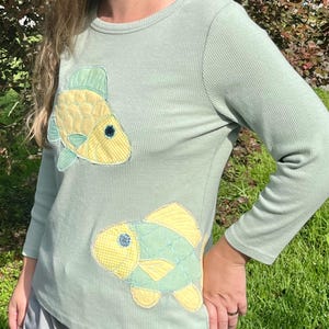 May include: A light green long-sleeve shirt with two fish appliqués. The fish are yellow and green with blue accents, and are made of patterned fabric. The shirt has a ribbed texture.