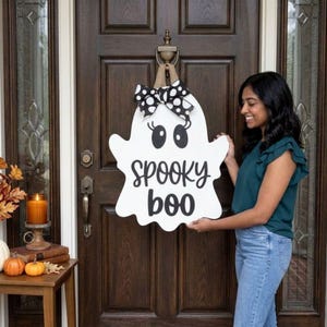 May include: A white wooden ghost-shaped door hanger featuring black eyes, eyelashes, and the text "spooky boo" in a black script font. The decoration is topped with a black and white polka dot fabric bow and a burlap hanging loop.