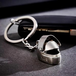 Può includere: Un portachiavi argentato a forma di casco, con il nome "Jack" inciso. Il portachiavi è attaccato a una catena e un anello argentati, appoggiato su una superficie scura.