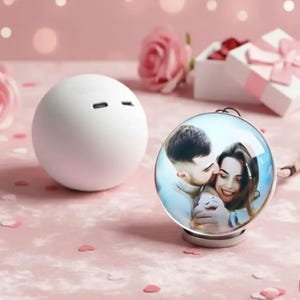 May include: A white spherical speaker with a smooth matte finish and two small ports on top, paired with a circular glass display stand featuring a printed photograph of a couple embracing, set against a soft pink background with scattered confetti.