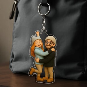 May include: A clear acrylic keyring depicting a cartoon couple in an embrace. The woman has red hair, a blue jacket, and a grey beanie. The man has a beard, glasses, and a beanie. The keyring hangs from a silver ring and chain.