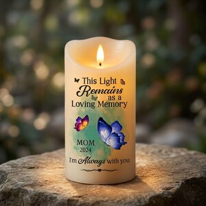 Può includere: Una candela LED cilindrica color crema con una fiamma luminosa. La candela presenta il testo "This Light Remains as a Loving Memory", "MOM", "2024" e "I'm Always with you", decorata con illustrazioni di farfalle colorate.