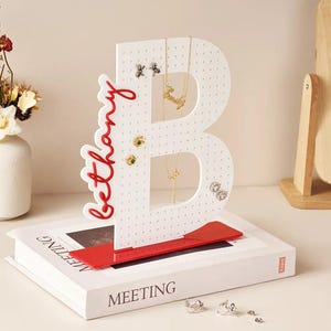 May include: A white, B-shaped jewelry organizer with small holes, holding necklaces and earrings. The name "Bethany" is written in red script on the side. The organizer sits on a white book with the word "MEETING" on the cover.