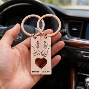 May include: Rose gold keychains with interlocking heart design. Each rectangular tag has a name and a date engraved. The keychains feature a heart-shaped cutout and a design of two hands holding pinky fingers.