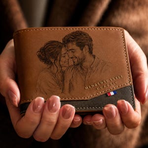 Puede incluir: Una cartera marrón de dos pliegues con un retrato grabado con láser de una pareja en la parte frontal. La cartera tiene una sección inferior de color marrón oscuro, costuras blancas y una pequeña etiqueta de tela roja, blanca y azul.
