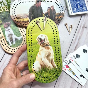 Peut inclure: Des planches de cribbage en bois avec des inserts photo personnalisés, représentant des images d'un chien, d'un couple et d'un chien. Les planches ont des motifs de points noirs et sont livrées avec des chevilles en bois. Un jeu de cartes est également présent.