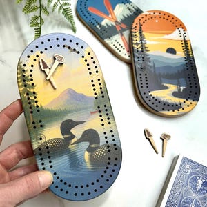 Peut inclure: Planches de jeu de cribbage en bois avec des motifs sur le thème de la nature. L'une représente un lac avec des huards, une autre une montagne et un coucher de soleil, et une troisième des skis. Chaque planche a des trous pour les chevilles et est livrée avec des chevilles en bois.