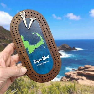 May include: Wooden cribbage board featuring a map of Cape Cod, Massachusetts, in green and blue. The board has a dark wood finish with drilled holes for pegs. Includes two white pegs with decorative tops. The text "Cape Cod - Massachusetts" is visible.