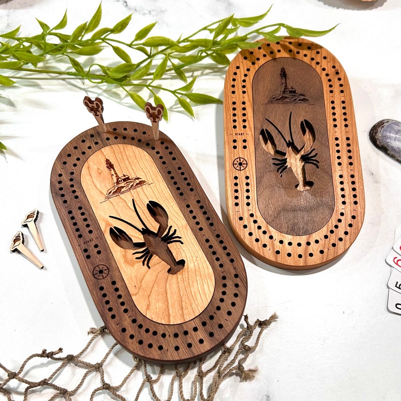 Peut inclure: Deux jeux de cribbage en bois avec un motif de homard et de phare. Les planches ovales ont un centre en bois clair et une bordure en bois fonc&eacute;. Des chevilles sont incluses.