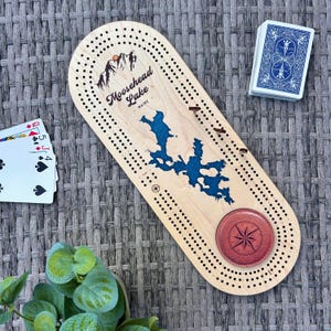 May include: A wooden cribbage board with the words "Moosehead Lake Maine" and a blue lake design. A red leather compass is at the bottom. A deck of cards and playing cards are next to the board.
