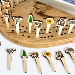Puede incluir: Un tablero de juego de madera con una variedad de clavijas de madera que presentan diferentes ilustraciones con temas de la naturaleza. Las clavijas incluyen un zorro, un colibrí, un árbol, un pez, un girasol, un búho y una furgoneta de camping. El tablero tiene un marco de madera y un patrón de cuadrícula negra.