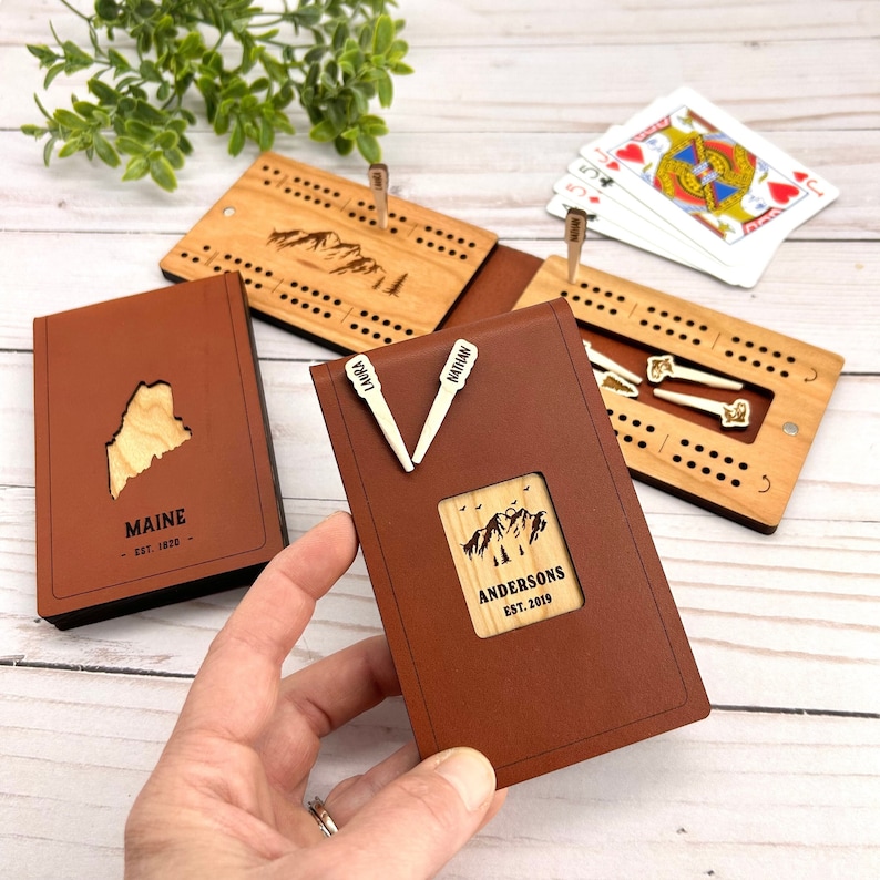 May include: A brown leather cribbage board with a wooden mountain scene and the text "ANDERSONS EST. 2019". The board is held by a hand. Other boards and a deck of cards are in the background.