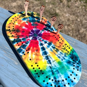 Peut inclure: Un jeu de cribbage tie-dye ovale avec des chevilles en bois. Les chevilles représentent des signes de paix et des gestes de la main. Le plateau présente un motif en spirale coloré en rouge, jaune, bleu et vert. Le plateau comporte de nombreux petits trous pour le placement des chevilles.