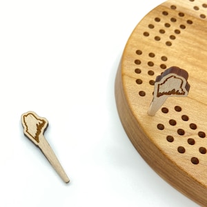 May include: A wooden game board with a circular shape and a grid of holes. Two wooden pegs with a state outline design are inserted into the board.