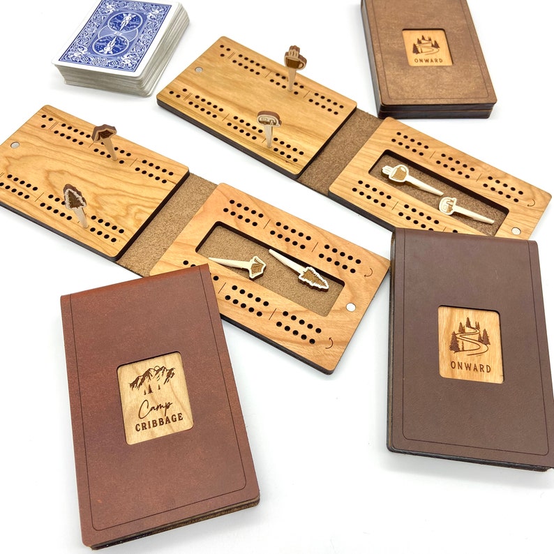 May include: A collection of wooden cribbage boards and card games. The boards are made of light-colored wood and feature peg holes and decorative wooden pegs. Some boards are open, revealing the interior. The covers are brown with the text "Camp Cribbage" and "Onward".