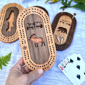 Peut inclure: Planches de cribbage en bois avec des motifs sur le th&egrave;me de la nature. L'une repr&eacute;sente un orignal, une autre un ours et une troisi&egrave;me un poisson. Chaque planche a une piste pour les chevilles et une rose des vents. Une main tient une planche, avec des cartes &agrave; jouer visibles.