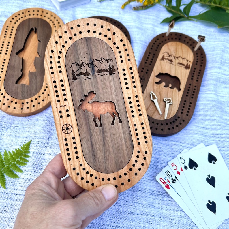 Peut inclure: Planches de cribbage en bois avec des motifs sur le th&egrave;me de la nature. L'une repr&eacute;sente un orignal, une autre un ours et une troisi&egrave;me un poisson. Chaque planche a une piste pour les chevilles et une rose des vents. Une main tient une planche, avec des cartes &agrave; jouer visibles.