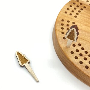 May include: A wooden peg game with two wooden pegs shaped like pine trees. The game board is made of light brown wood and has a circular shape. The pegs are inserted into holes in the board.