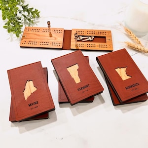 May include: A wooden cribbage board with a brown leather case and state-shaped cutouts. The board is open, showing pegs and scoring holes. Several brown leather cases with state outlines are stacked nearby. The state names Maine, Vermont, and Minnesota are visible.